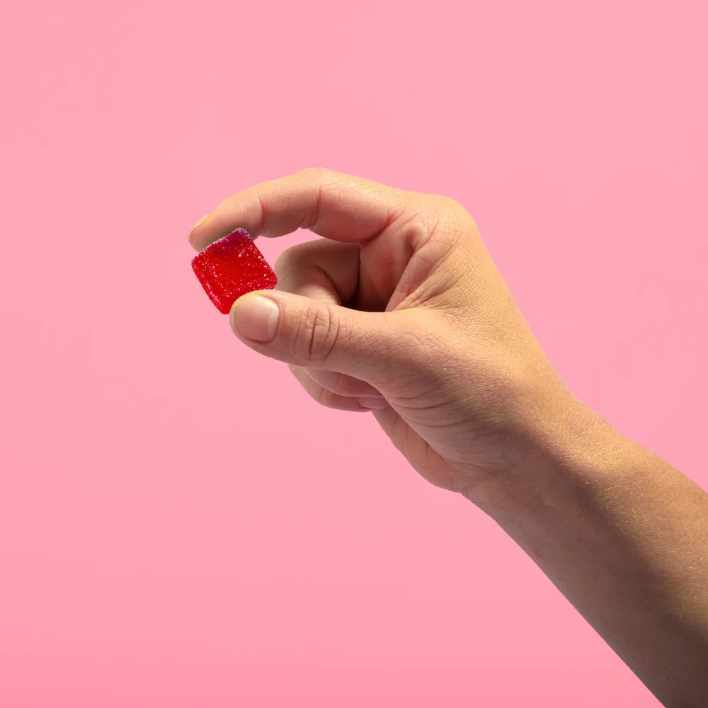 A hand holding a red gummy candy against a pink background.