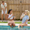 Two women sitting by a pool, one holding a product, while a man plays golf in the background.